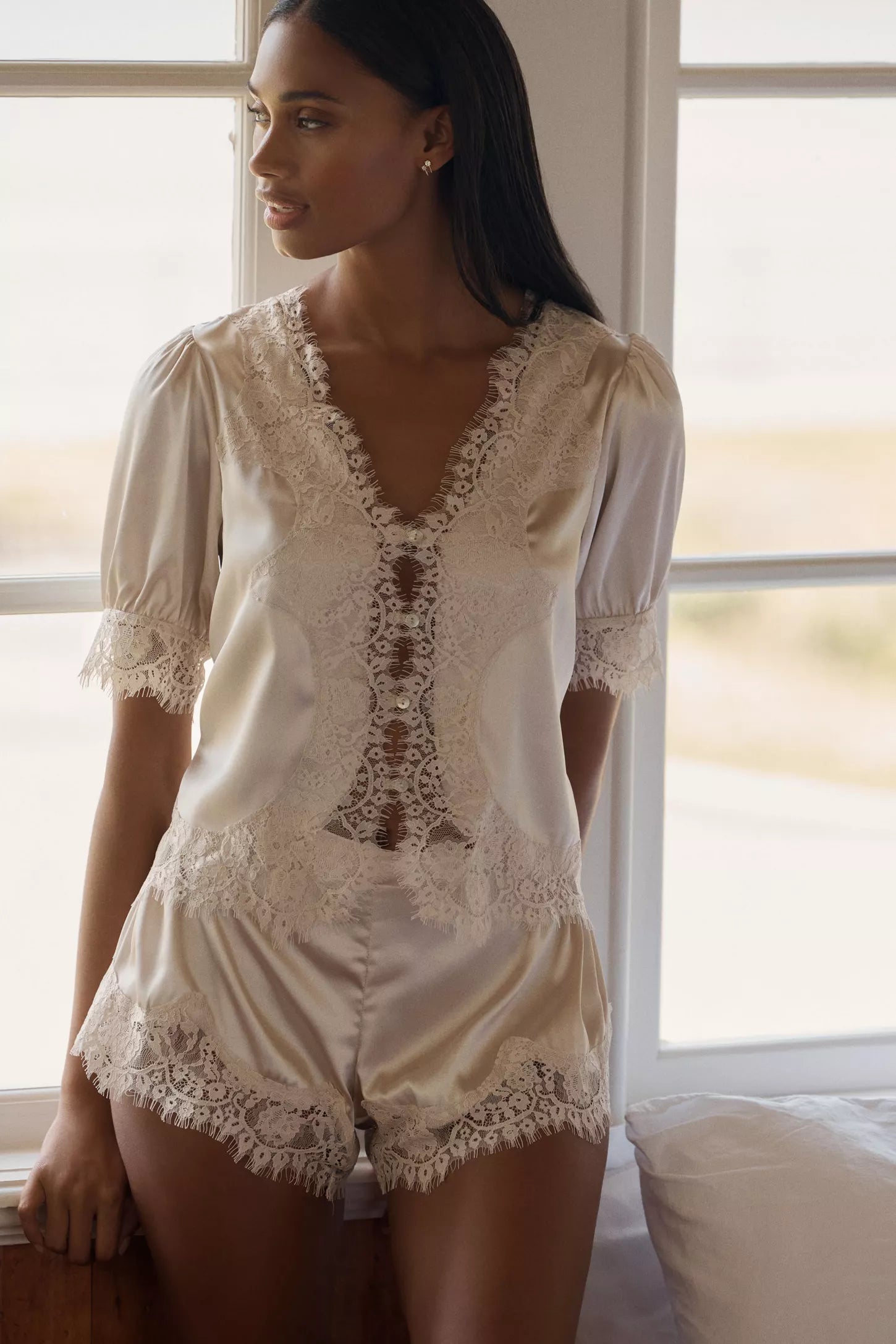 A woman wearing a cream satin short-sleeve lingerie top and matching lace-trimmed shorts, posed in front of a bright window.